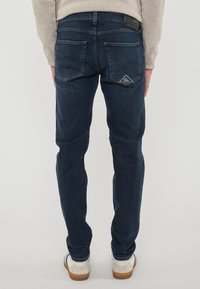 Persona che indossa jeans in denim blu scuro con tasche posteriori e scarpe da ginnastica bianche, in piedi con le braccia rilassate ai lati.