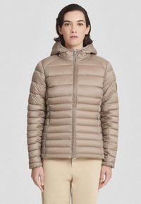 Vrouw in een lichtbruine gewatteerde jas met capuchon, dichtgeritst, en beige broek, staand tegen een lichte effen achtergrond.