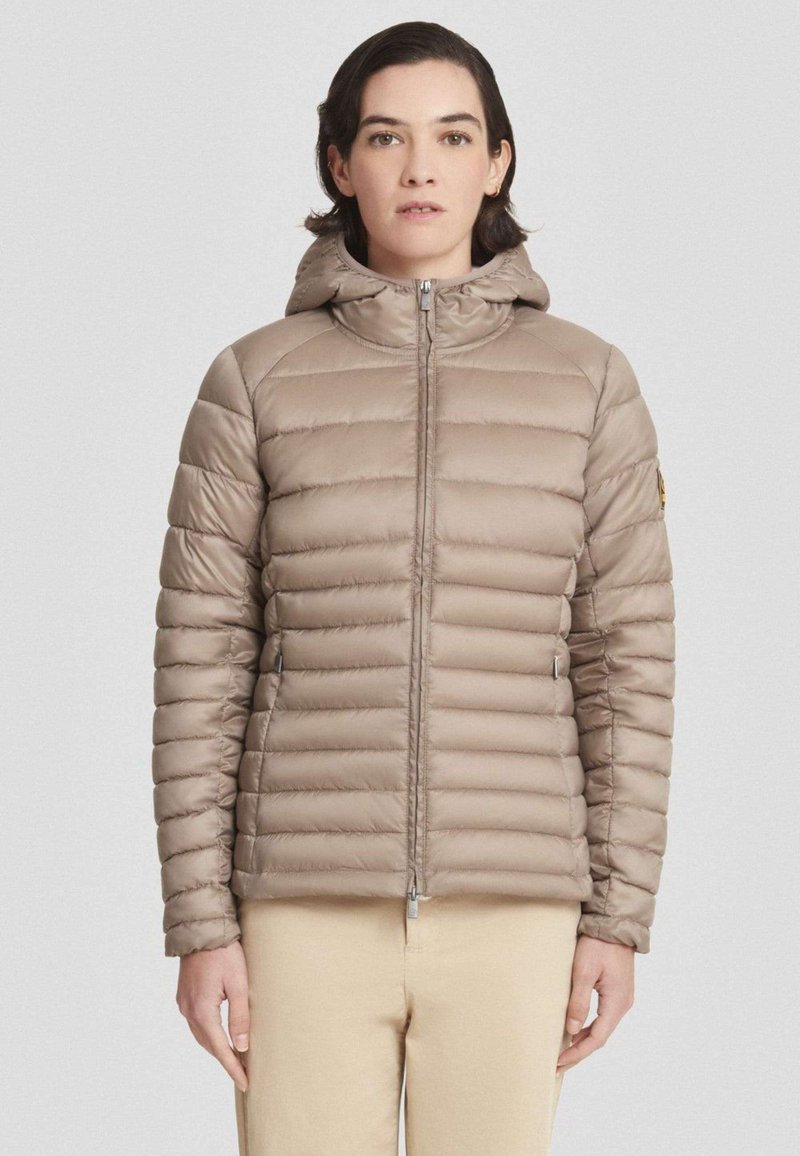 Vrouw in een lichtbruine gewatteerde jas met capuchon, dichtgeritst, en beige broek, staand tegen een lichte effen achtergrond.