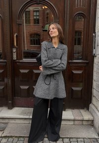 Sivý vlnený kabátový šaty s golierom, s gombíkovým zapínaním a viazaním v páse. V kombinácii so širokými čiernymi nohavicami. Pozadie z dreva.