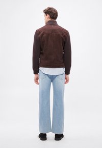 Jachetă maro din piele întoarsă și tricot cu guler înalt, asortată cu blugi largi de culoare albastru deschis și pantofi negri, văzută din spate.