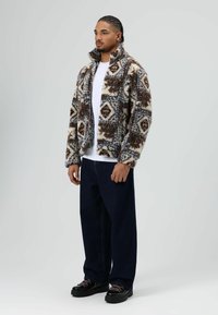 Nepbonten jas met geometrische patronen in bruin, wit en blauw. Opstaande kraag, lange mouwen. Gecombineerd met losse donkerblauwe spijkerbroek en zwarte schoenen.