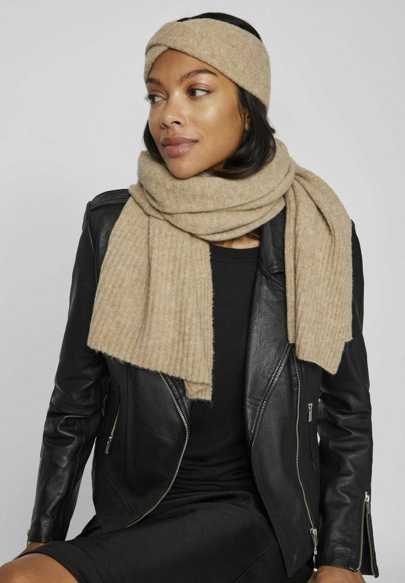 Sciarpa a coste beige con fascia abbinata, realizzata in morbida lana, indossata sopra una giacca di pelle nera, che accentua un look casual.