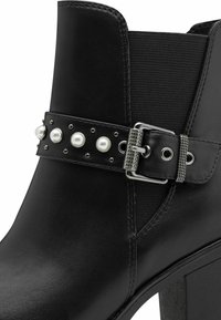Black ankle boot featuring a smooth leather finish, elastic panel, and decorative strap with pearls and studs, secured by a metallic buckle.
