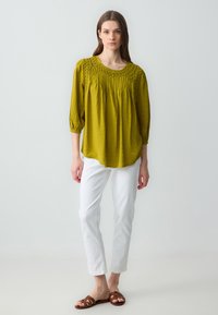 Blusa verde oliva con texture plissettata e collo ricamato, abbinata a pantaloni bianchi e sandali marroni con cinturini.