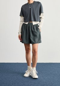 Šedé zkrácené sportovní tričko vrstvené přes bílé tričko s dlouhým rukávem, doplněné o zelené šortky a bílé vysoké tenisky.