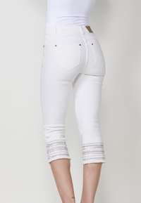 Jean capri blancs avec une coupe ajustée. Présente un ourlet effiloché et un détail texturé en rayures au niveau du poignet. Deux poches arrière avec des coutures décoratives.