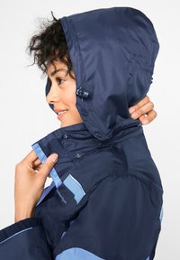 Giacca impermeabile blu navy con cappuccio, caratterizzata da dettagli blu chiaro, chiusura con zip e bottoni sul colletto e sulle maniche.