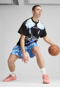 Sort grafisk t-shirt med blå vinge designs, parret med blå mønstrede shorts. Personen bærer ferskenfarvede sportsko, mens han dribler med en basketball.