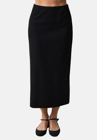Jupe noire mi-longue avec une coupe droite et une texture minimale. Elle présente une taille lisse et est associée à des ballerines noires. Aucune motif visible.