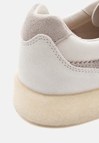 Sapatilha cinza-claro com um cabedal de couro liso e discretos toques de camurça cinza. Apresenta uma sola texturizada e costuras ao longo das bordas.