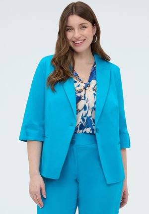 Donna sorridente che indossa un blazer azzurro acceso con le maniche arrotolate, pantaloni abbinati e una camicetta con motivo floreale con accenti blu e marroni.