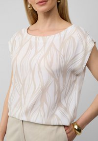 Witte blouse met een lichtbeige abstract bladpatroon, ronde halslijn, korte mouwen en een gladde stof. Gouden armband om de pols.