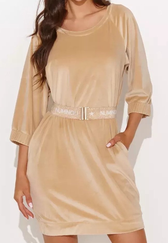 Femme portant une robe en velours beige avec des manches trois-quarts et une ceinture scintillante, une main dans la poche, debout devant un fond blanc.