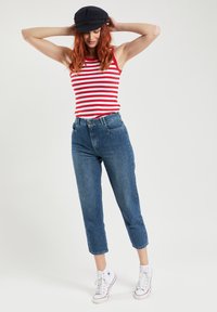 Rood en wit gestreepte tanktop, high-waisted blauwe spijkerbroek en witte sneakers. Model draagt een zwarte pet en staat met de handen op het hoofd.