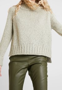 Ljusgrön stickad tröja med tjock ribbad nederkant och ärmar. Har en hög halsringning och avslappnad passform, bärs över mörkgröna läderbyxor.