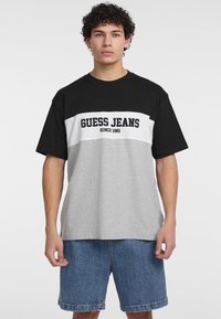 Juoda ir pilka trumpomis rankovėmis marškinėliai su baltu paneliu ir užrašu "GUESS JEANS SINCE 1981". Minkšta medžiaga, laisvas kirpimas ir apvalus iškirpimas.
