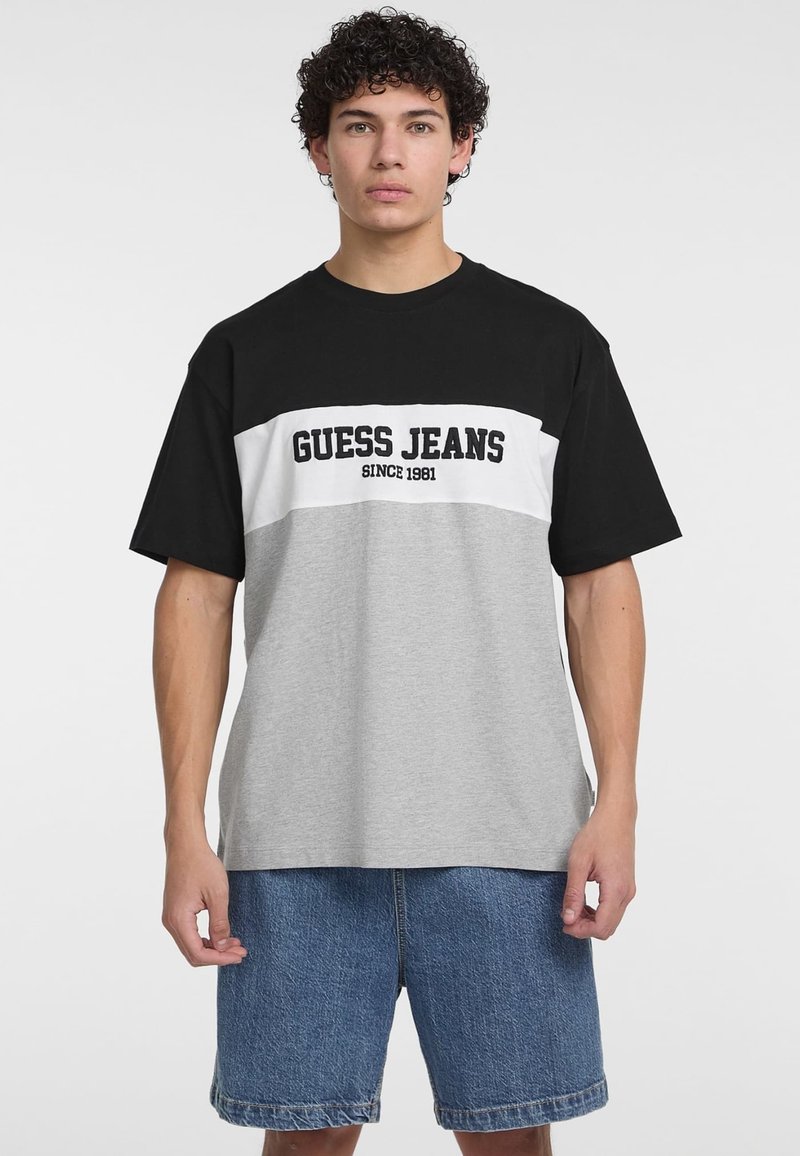 Juoda ir pilka trumpomis rankovėmis marškinėliai su baltu paneliu ir užrašu "GUESS JEANS SINCE 1981". Minkšta medžiaga, laisvas kirpimas ir apvalus iškirpimas.