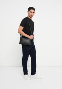 Μαύρη δερμάτινη τσάντα messenger με υφή, επάνω φερμουάρ και ρυθμιζόμενο λουρί, συνδυασμένη με ναυτικές παντελόνες και λευκά αθλητικά παπούτσια.