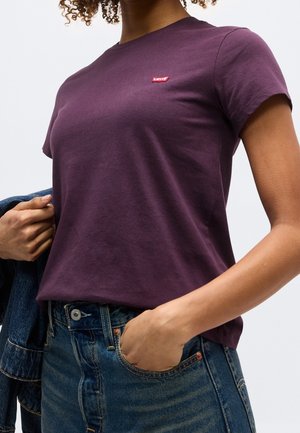 Nainen pukeutunut violettiin Levi's T-paitaan ja sinisiin farkkuihin, pitää farkkutakkia toisella kädellä ja laittaa toisen käden taskuun.