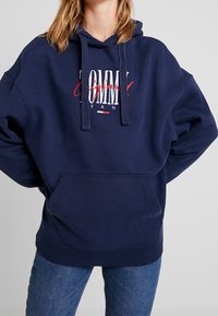 Marinblå hoodie med snören, stor framficka och vit/röd "TOMMY" logotekst. Tillverkad av ett mjukt, texturerat tyg.