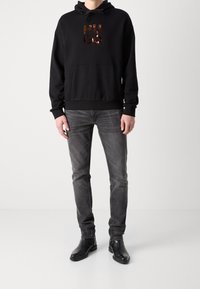 Sudadera negra con un logotipo metálico de "HUGO", bolsillo canguro, combinada con jeans grises y botas de cuero negro pulido. Diseño simple y casual.