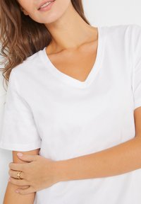 T-shirt bianco a V con maniche corte, caratterizzato da una texture morbida e un fit comodo. La mano del modello poggia sul braccio, indossando un anello d'oro.