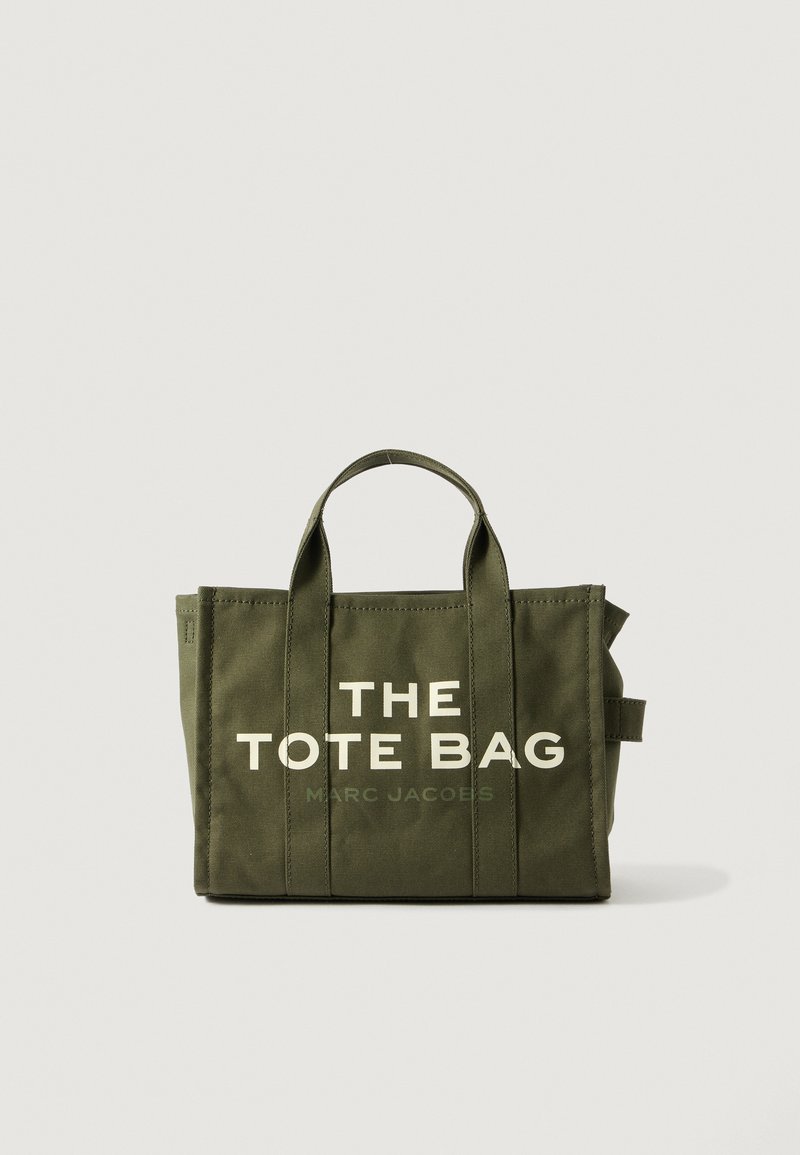 Green canvas tote bag with double handles, featuring bold white text: "THE TOTE BAG" and "MARC JACOBS." Simple, rectangular design.