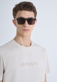 Gafas de sol negras con forma angular, llevadas por un modelo. Se presenta una camiseta de color beige claro con un sutil logo "MORATO" en relieve en el pecho.