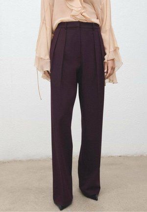 Kobieta w ciemnofioletowych, szerokich spodniach z wysokim stanem, do których dobrała przezroczystą beżową bluzkę z marszczonymi rękawami, stojąca na gładkiej podłodze.