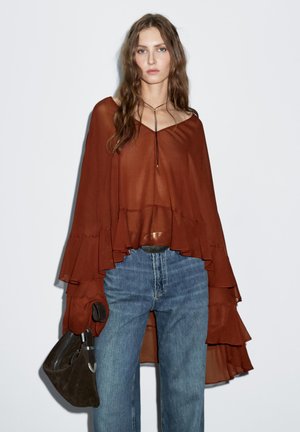 Brown layered blouse with sheer fabric, wide sleeves, and a V-neck. Paired with light blue straight-leg jeans and a dark handbag.