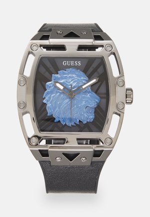 Montre carrée en argent avec bracelet noir, motif tête de lion bleu sur cadran noir, aiguilles argentées, et logo "GUESS" en haut au centre.