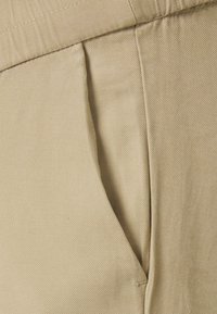 Calças de algodão bege com um acabamento texturizado, apresentando um bolso lateral e pregas estruturadas na área da cintura. Design simples e funcional.