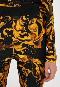 Black fabric featuring gold baroque-style patterns. The waistband is elastic with embossed text. Close-up of a decorative design with intricate swirls.