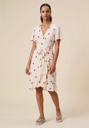 Femme portant une robe portefeuille crème avec un imprimé floral rouge et des manches volantées, nouée à la taille, debout dans une posture détendue avec un fond neutre.