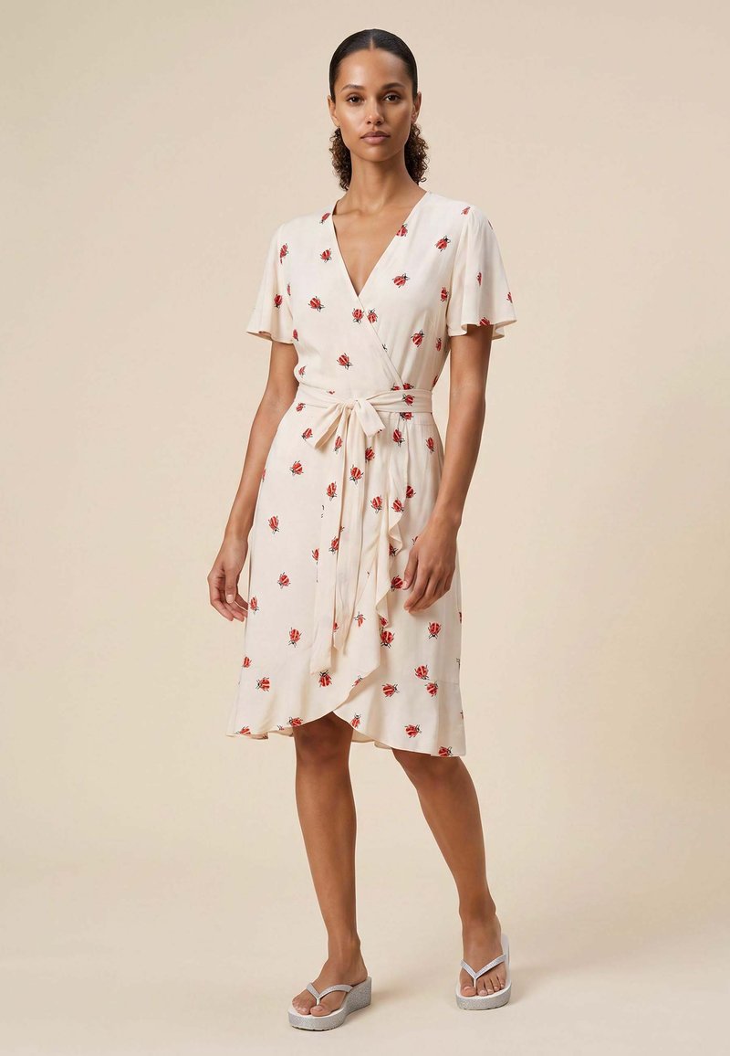 Femme portant une robe portefeuille crème avec un imprimé floral rouge et des manches volantées, nouée à la taille, debout dans une posture détendue avec un fond neutre.