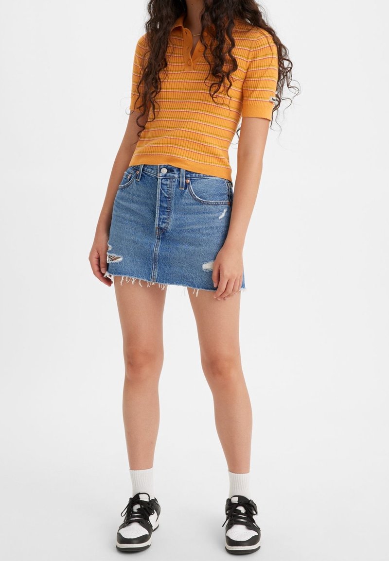 Femme portant un polo côtelé orange à manches courtes, une mini-jupe en jean bleue délavée, des chaussettes blanches et des baskets noires et blanches, debout sur fond blanc.