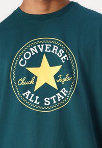 Camiseta de algodón en color teal con un gráfico circular amarillo que presenta una estrella, y texto en blanco que dice "Converse," "All Star" y "Chuck Taylor."