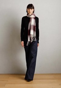 Svart langermet topp kombinert med vide mørke denimbukser. Et mønstret skjerf har krem, burgunder og grå nyanser med frynser.