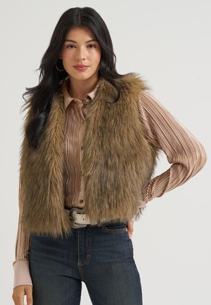 Femme aux longs cheveux foncés porte un gilet en fausse fourrure marron sur un chemisier en satin plissé et un jean foncé avec une ceinture blanche, debout devant un fond uni.