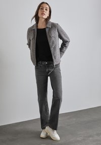 Veste grise texturée avec un col, pull noir en dessous, jean gris et baskets blanches. Pose debout contre un fond uni.