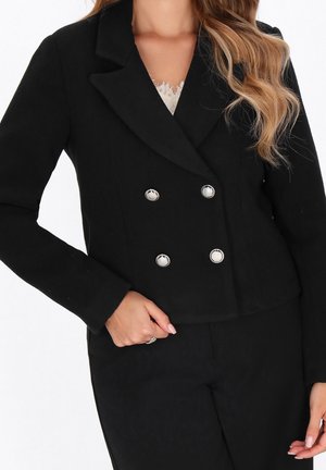 Vrouw draagt een zwarte dubbelgeknoopte blazer met zilveren knopen over een wit topje met kanten randje, hand rust bij de taille.