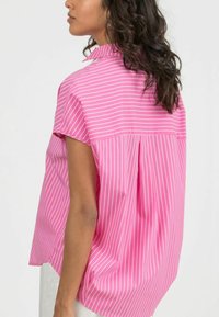 Blusa rosa a maniche corte con righe verticali bianche, caratterizzata da un colletto alto, vestibilità morbida, piega sul retro e texture di tessuto liscia.