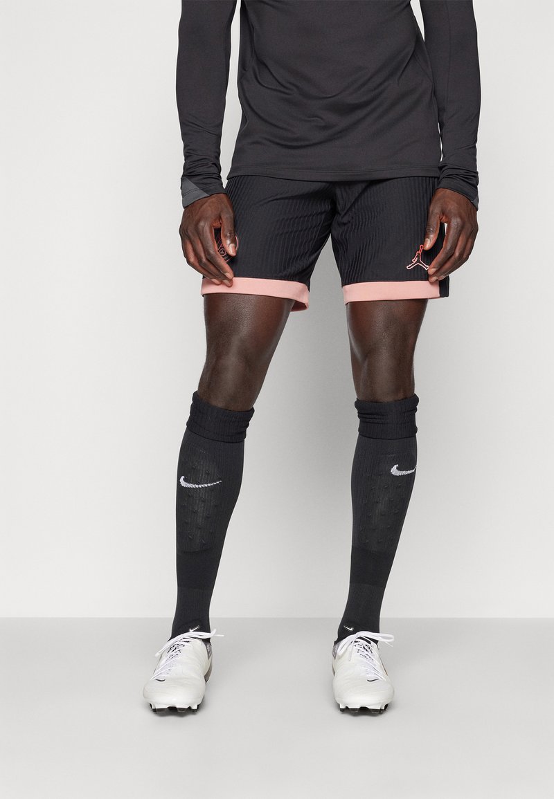 Schwarzes langärmliges Oberteil, schwarze Shorts mit pinken Bündchen, schwarze Fußballsocken mit Nike-Logo und weiße Fußballschuhe. Strukturierte Stofftextur.