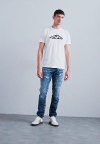 White cotton T-shirt with black logo, paired with distressed blue denim jeans. Model wears white sneakers with black stripes.