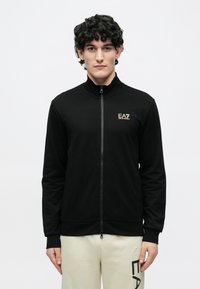Čierna bunda na zips s vysokým golierom, s logom EA7 v zlatej farbe na ľavej strane hrudníka. Hladký materiál, elastické manžety a pás.