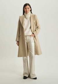 Manteau en laine beige avec un grand col, pull col roulé crème, et pantalons argentés évasés brillants. Le mannequin se tient contre un fond neutre.