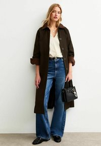 Abrigo trench de button-up marrón con un collar en contraste, blusa blanca, jeans anchos azules y un pequeño bolso negro con accesorios metálicos.