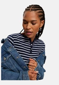 Granatowy i biały pasiasty top z długim rękawem z guzikami, w połączeniu z wyblakłą kurtką dżinsową z widocznym przeszyciem i metalowymi detalami.