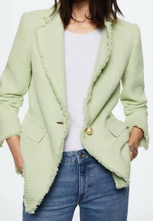 Mujer con blazer verde claro texturizado con bordes deshilachados, camiseta blanca y vaqueros azules, manos en los bolsillos, mostrando un tatuaje en la muñeca.